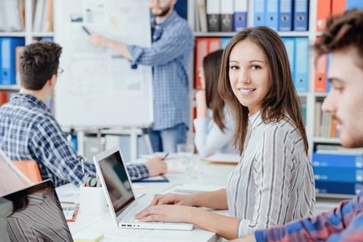 People sitting in a meeting on a table with laptops in front of them and a person in the background writing something on a flipchart